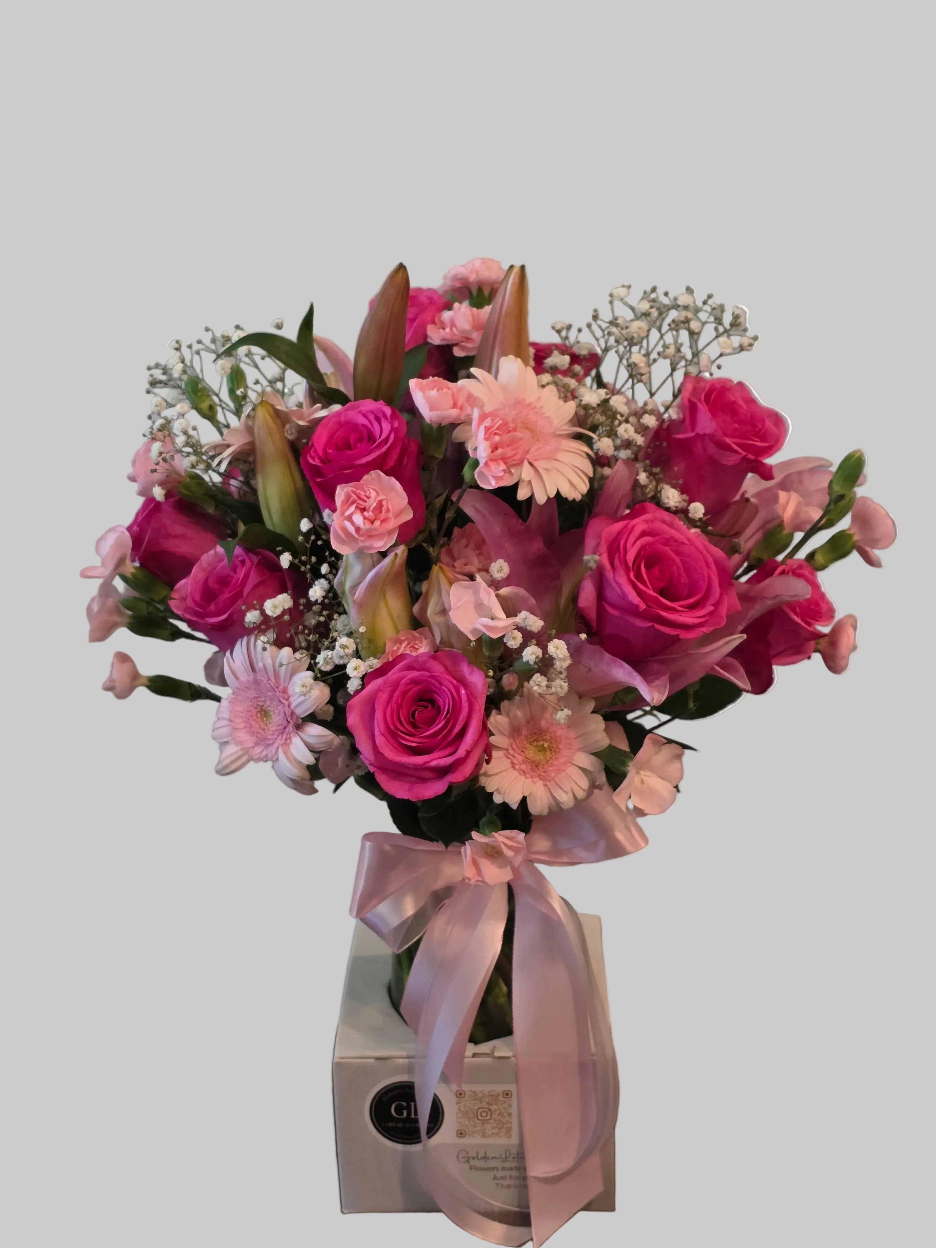 Pink floral arrangement with roses, lilies, gerbera daisies, carnations, and baby's breath in a glass vase with a pink ribbon.