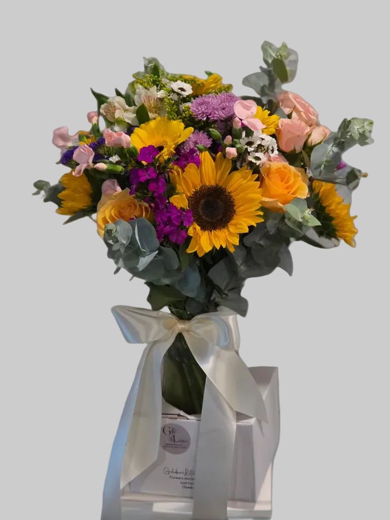 Bright sunflower bouquet with yellow roses, pink spray roses, chrysanthemums, and eucalyptus arranged in a glass vase with satin ribbon.